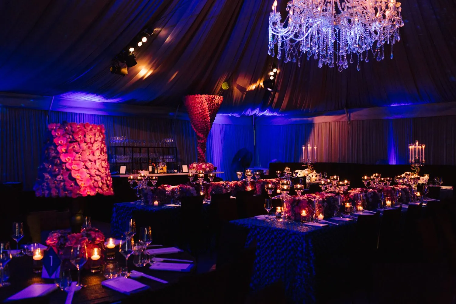 Draped holiday tent with crystal chandeliers, candlelit tables, and deep blue uplighting