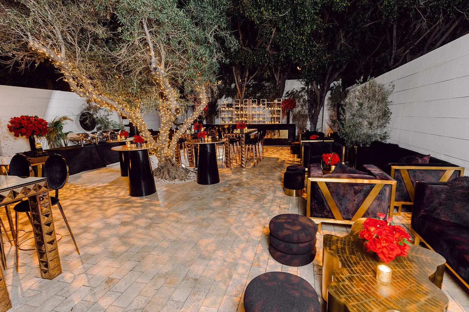 Private party courtyard lounge with a twinkle-wrapped olive tree casting dappled light over gold and black furnishings